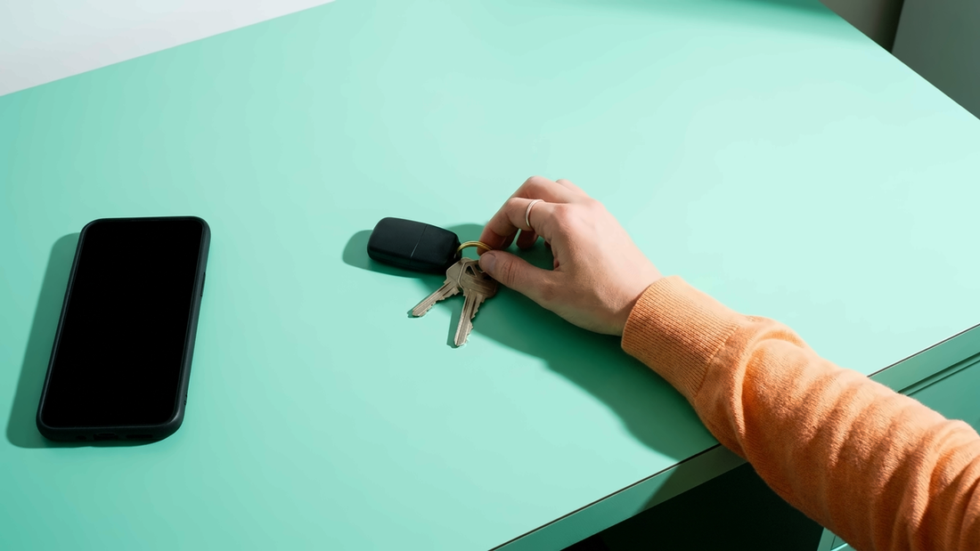 A hand in an orange sleeve picks up a set of car keys from a mint-green desk in cool daylight, a phone lying face-up and dark beside them.