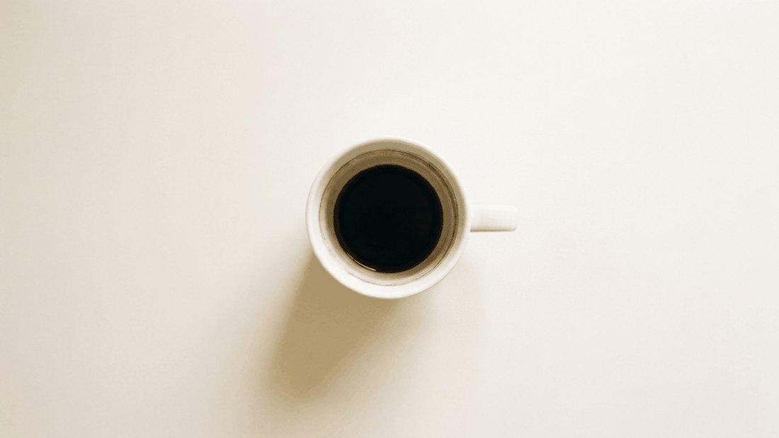 A single white cup of black coffee photographed from directly above on a plain off-white surface in flat daylight.