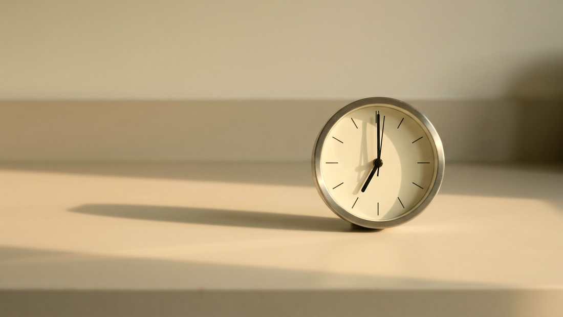 A small silver kitchen clock reading 7:00, alone on an empty pale kitchen counter in low warm evening light.
