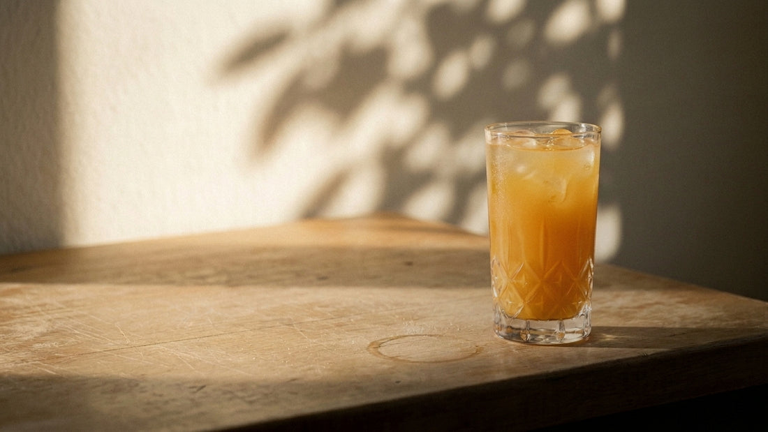 A single glass of Third Nature peach-mango drink on a worn wooden kitchen counter in low early-September morning light, with dappled leaf shadow on the wall behind.